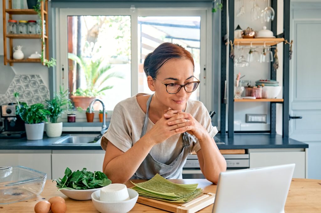 Woman preparing homemade lasagna with spinach and ricotta from recipe found online in her laptop.
